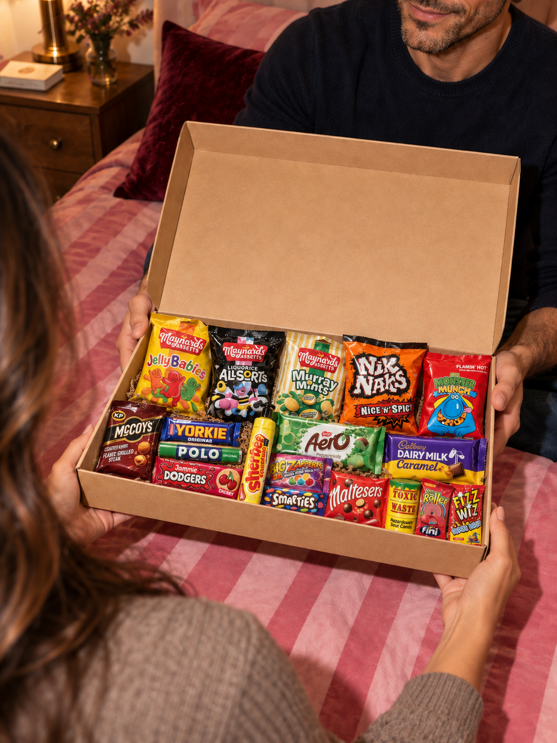 A couple sharing an example HRS Snack Club gift box with sweets, chocolate and crisps. Products and pack sizes vary.